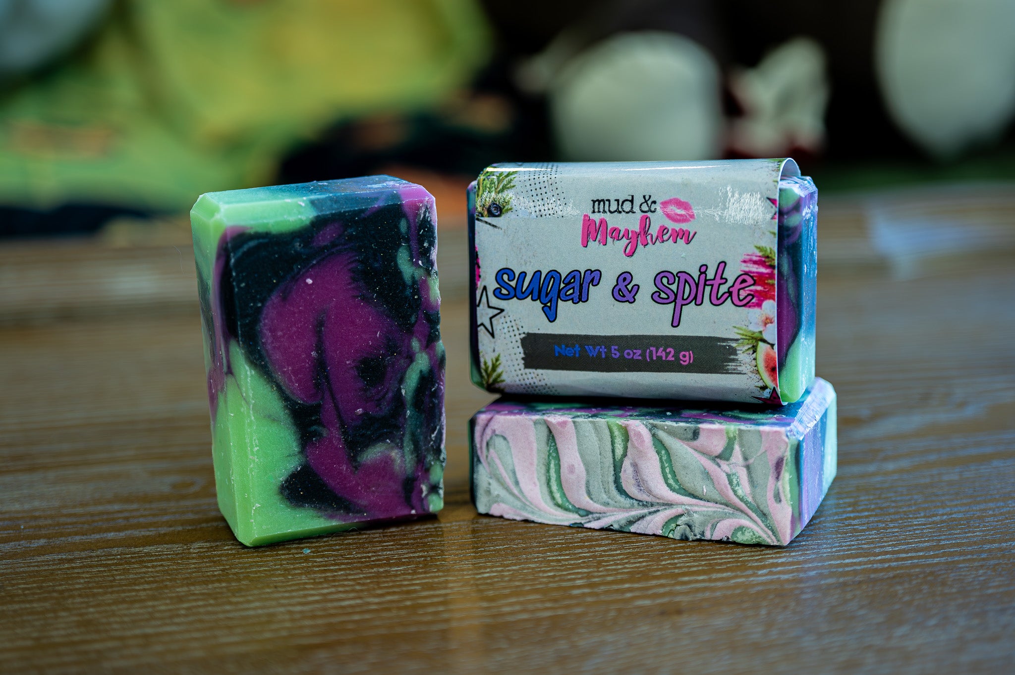 Three handmade marbled soap bars and one wrapped soap bar on a wooden table. The soaps feature swirls of green, purple, black, pink, and white.