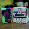 Three handmade marbled soap bars and one wrapped soap bar on a wooden table. The soaps feature swirls of green, purple, black, pink, and white.